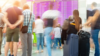Viele Personen in Bewegungsunschärfe mit und ohne Koffern stehen vor einer große, elektronischen Anzeigetafel, auf der Information steht. Auf der Anzeigetafel steht: Departures sowie Uhrzeiten, Nummern und Städte.