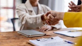 Zwei Personen sitzen sich an einem Tisch gegenüber und schütteln sich die Hände. Vor einer Person liegt ein Zettel, auf dem Vertrag steht. Daneben steht eine weitere Person, die applaudiert