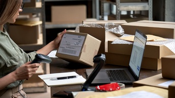 Person mit Brille scannt Kartonagen in einem Lagerraum, daneben stehen ein Laptop sowie ein Notizbuch mit einem Stift und Verpackungsmaterialien, im Hintergrund stehen Regale mit Kartonagen