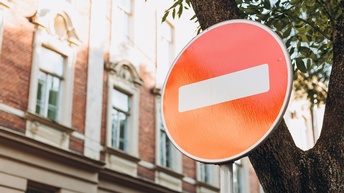 Detailansicht eines Straßenschildes für Fahrverbot: Kreisrundes rotes Schild mit weißem Streifen mittig, im Hintergrund Ast eines Baumes und verschwommen Hausfassade
