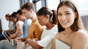 Jugendliche Person mit langen braunen Haaren sitzt gemeinsam mit anderen Jugendlichen wartend bei einer Fensterfront