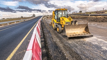 Ein gelber Bagger mit Frontschaufel steht auf einer unbefestigten Fläche neben einer asphaltierten Straße. Links verläuft die Fahrbahn mit Leitplanke und rot-weißen Absperrelementen. Rechts befindet sich ein Baustellenbereich mit Erde und Schotter