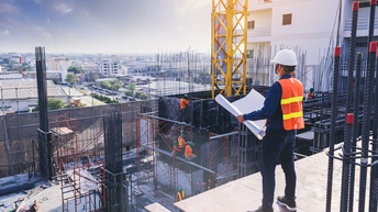 Eine Person in Sicherheitsweste und Helm steht auf einer erhöhten Plattform und blickt in den Bildhintergrund. In den Händen hält sie einen großen, weißen Zettel. Unter der Plattform ragen viele Metallstäbe aus dem betonierten Boden.