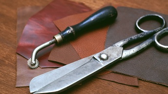 Nahaufnahme einer silbernen Schere und eines Werkzeugs mit einem kleinen gezackten Rad am Ende, die auf kleinen Stoffmustern sind. Die Stoffmuster haben unterschiedliche Brauntöne