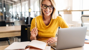 Lächelnde Person mit Brille und erhobenem Stift in der Hand sitzt vor aufgeklapptem Laptop und Büchern 