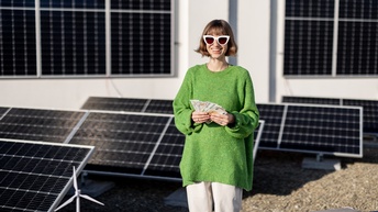 Lächelnde Person in grünem Pullover mit weißen Sonnenbrillen steht inmitten einer Solaranlage und hält Fächer mit Eurogeldscheinen in Händen, im Vordergrund Modell eines Windrades