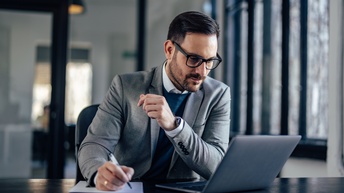 Person mit kurzen Haaren, Brille und Bart trägt Businesskleidung und arbeitet bei einem Schreibtisch mit einem Laptop in einem Büro