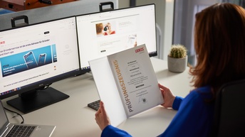 Person mit langen braunen Haaren und blauer Bluse sitzt an einem Schreibtisch mit Laptop und zwei Monitoren und hält ein Dokument mit Aufschrift Prüfungszeugnis Meisterin in Händen