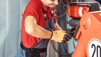 Person in Arbeitslatzhose mit Handschuhen schraubt mit Werkzeug an orangem Gefährt, das im Ausschnitt erkennbar ist