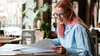 Person mit langen rosaroten Haaren und Brille blickt lächelnd auf Papierdokumente