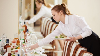 Person in weißer Bluse beugt sich über gedeckten Tisch und greift nach Schüssel mit einer Hand, in anderer Hand hält sie Teller mit Essen