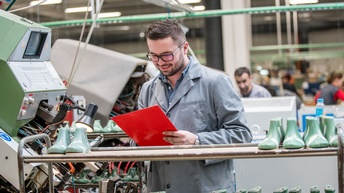 Person mit Brillen blickt auf rote Mappe in Händen, ringsum Schuhmodelle in Industriehalle