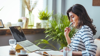 Seitliche Aufnahme einer an einem Schreibtisch sitzenden Person. Vor ihr ist ein aufgeklapptes Buch, auf das sie blickt. Auf dem Tisch sind ein Laptop, eine Tasse, ein Tablet, eine Sanduhr und ein Glas. Im Hintergrund sind Topfpflanzen