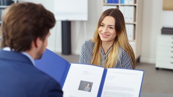 Eine Person mit langen, blonden, offenen Haaren sitzt einer zweiten Person mit dunklen, kurzen Haaren gegenüber, die den Lebenslauf und ein Schreiben der erstgenannten Person in den Händen hält.