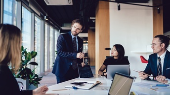 Um einen Tisch sitzen drei Personen. Am Tisch steht eine Person, die einen Zettel mit einer Grafik hält. Die anderen Personen blicken zu der stehenden Person. Auf dem Tisch sind Laptops, Stifte, Notizbücher sowie verschiedene Zettel und Unterlagen