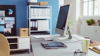 Arbeitsplatz in Büro mit Computerbildschirm und diversen Arbeitsutensilien, im Hintergrund Regal mit Aktenordnern