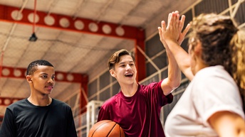 Drei Personen in Sporthalle, zwei Personen schlagen die Hände zum High Five aneinander, eine der Personen hält in anderer Hand Basketball