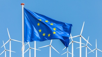 EU-Flagge im Wind wehend, im Hintergrund Windräder unter blauem Himmel