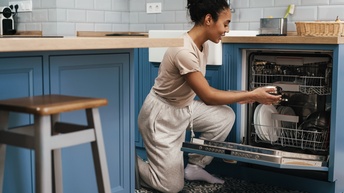 Person hockt vor offenem Geschirrspüler in Küche mit blauen Fronten und hält Pfanne in Händen