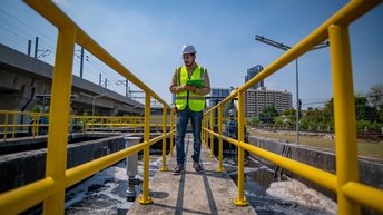 Person in gelber Warnweste mit weißem Helm geht über mit gelbem Geländer eingefassten Steg einer Wasseraufbereitungsanlage, links und rechts vom Steg umwälzendes Wasser unter blauem Himmel