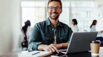 Nahaufnahme einer Person mit Brille, die vor einem aufgeklappten Laptop sitzt, der auf einem braunen Bürotisch steht. Am Tisch ist außerdem ein Coffee-to-go-Behälter, ein Smartphone und ein Notizblock mit einem Stift