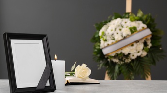 Schwarzer Stahbilderrahmen mit schwarzem Band auf Tisch stehend, daneben brennende weiße Kerze und aufgeschlagenes Buch mit weißer Rose, im Hintergrund verschwommen weißgrünes Blumenbouquet mit Scherpe