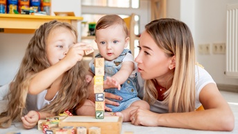 In der Mitte sitzt ein Kleinkind. Links daneben liegt ein Kind auf dem Bauch. Rechts neben dem Kleinkind ist eine erwachsene Person, die das Kleinkind mit dem rechten Arm umfasst. Vor ihnen steht ein Turm aus Holzwürfeln