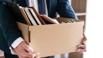 Nahaufnahme eines kleinen, rechteckigen Kartons, der von den Händen einer Person in einem Anzug gehalten wird. In dem Karton sind Bücher, Notizhefte und ein Klemmbrett vertikal nebeneinander angeordnet.