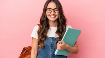 Porträt einer lächelnden Person mit offenen, dunklen, langen Haaren, Brille Jeanskleid, Notizbüchern und Rucksack, die vor einer rosa Wand steht