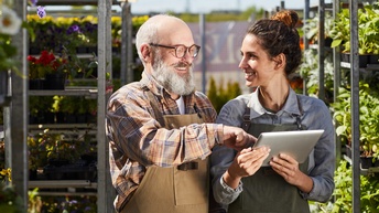 Nahaufnahme von zwei Personen, die in einer Gärtnerei nebeneinander stehen und sich ansehen. Die rechte Person hat einen Bart und eine Brille und deutet auf das Tablet, das die linke Person in den Händen hält. Im Hintergrund Metallregale mit Pflanzen