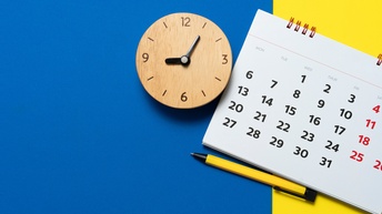 Auf blau-gelbem Hintergrund liegt eine Uhr aus Holz. Neben der Uhr liegt ein Kalenderblatt eines Ringbuchkalenders, unter dem ein Kugelschreiber liegt