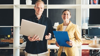 Zwei stehende Personen mit Laptop und Clipboard in Händen, im Hintergrund Büroraum