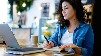 Person blickt auf aufgeklapptes Notebooks und nimmt mit Stift Notizen in Buch vor