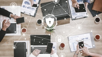Top-Shot einer hölzernen Oberfläche, auf der aufgeklappte Laptops, Klemmbretter mit Papieren mit Statistiken, Tassen mit Flüssigkeiten, die Hände von Personen mit Smartphones sind. Darüber sind kleine Schlösser umgeben von Kreisen