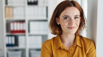 Portrait einer Person mit schulterlangen rötlichen Haaren und Perlohrstecker sowie gelben Poloshirt steht bei einer Wand, im Hintergrund zeigt sich ein Regal mit Mappen