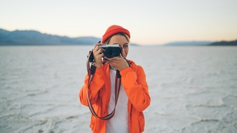 Person in oranger Jacke mit oranger Haube in Wüstenlandschaft stehend lugt durch Sucher einer Fotokamera