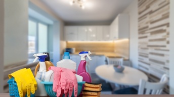 Im Vordergrund sind auf einem Holztisch verschiedene Utensilien zum Putzen, zum Beispiel Gummihandschuhe, Sprühflaschen, Putzschwämme. Im Hintergrund ist eine Küche mit einem Tisch