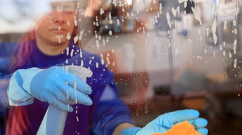 Aufnahme einer Person mit Handschuhen durch eine Glasscheibe, die in der linken Hand ein Tuch hält und in der rechten eine Sprühflasche. Auf der Glasscheibe ist bedeckt mit einer schäumenden Flüssigkeit.