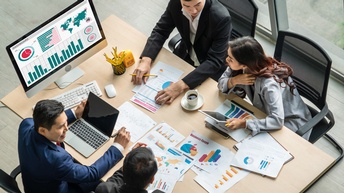 Personen in Businesskleidung sitzen an einem Tisch und besprechen Analysen in Form von ausgedruckten Unterlagen, am Laptop sowie auf einem Computerbildschirm ersichtlich, Topview