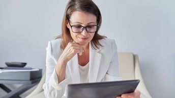 Eine Person in einem hellen Sakko, mit offenen, braunen Haaren und Brille sitzt auf einem cremefarbenen Sessel und hält ein Tablet, auf das sie blickt