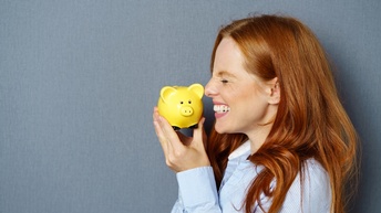 Eine Person mit langen orangenen Haaren steht seitlich vor einer grauen Wand und hält lachend ein gelbes Sparschwein in den Händen.