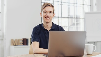 Porträt einer an einem Tisch sitzenden Person, die vor sich einen aufgeklappten Laptop hat. Neben dem Laptop sind ein Notizbuch und ein Stift