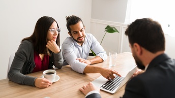 An einem Tisch sitzt eine Person in einem Sakko. Der Person gegenüber sitzen zwei Personen. Zwischen ihnen steht ein aufgeklappter Laptop, auf den eine Person deutet. Die andere Person hat eine Tasse Kaffee vor sich