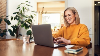 Eine Person mit Brille, offenen, hellen Haaren und Pullover sitzt an einem runden Tisch in einem Wohnzimmer und tippt auf der Tastatur eines Laptops. Im Hintergrund sind Zimmerpflanzen und ein Fenster