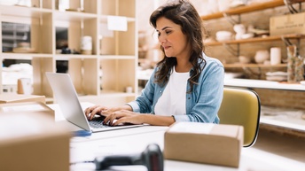 Eine Person sitzt hinter einem Tisch. Auf dem Tisch sind ein paar kleine Kartons. Die Person blickt auf den Bildschirm eines Laptops, der vor ihr steht. Hinter der Person ist ein Regal mit unterschiedlichen Gegenständen