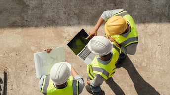 Topshot dreier Personen mit Helmen und Sicherheitswesten, die über eine Baustelle gehen. Die Person links hält einen Plan, die Person in der Mitte einen Laptop und die Person rechts deutet nach vorne mit dem Zeigefinger
