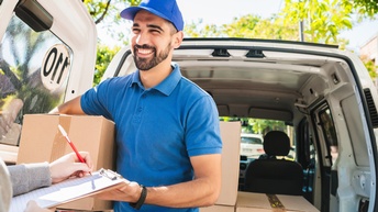 Eine Person in blauem Poloshirt und Kappe hält ein Paket vor einem geöffneten Lieferfahrzeug. Im Vordergrund schreibt eine weitere Person mit einem Stift auf ein Klemmbrett. Im Laderaum des Fahrzeugs befinden sich weitere Kartons