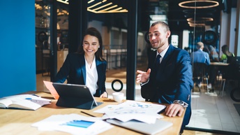 Zwei Personen in dunklen Anzügen sitzen in Besprechungszimmer vor Tablet, am Tisch liegen Unterlagen