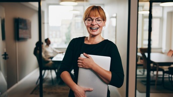 Lachende Person mit Brillen hält Notebook in Armen, im Hintergrund verschwommen Besprechungszimmer mit sitzenden Personen