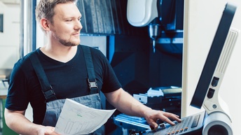 Person in Arbeitskleidung hält einen Plan in der Hand und bedient motiviert eine CNC Maschine